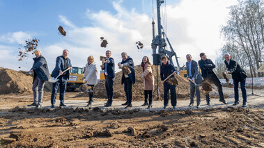 Gruppe von Personen mit Spaten bei einem symbolischen Spatenstich auf einem freien Gelände.